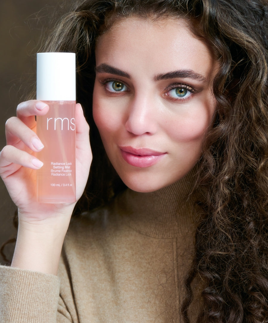 Woman with curly hair holding a bottle of Radiance Lock Setting Mist by RMS Beauty, showcasing clean beauty and skincare infused makeup in sustainable packaging.