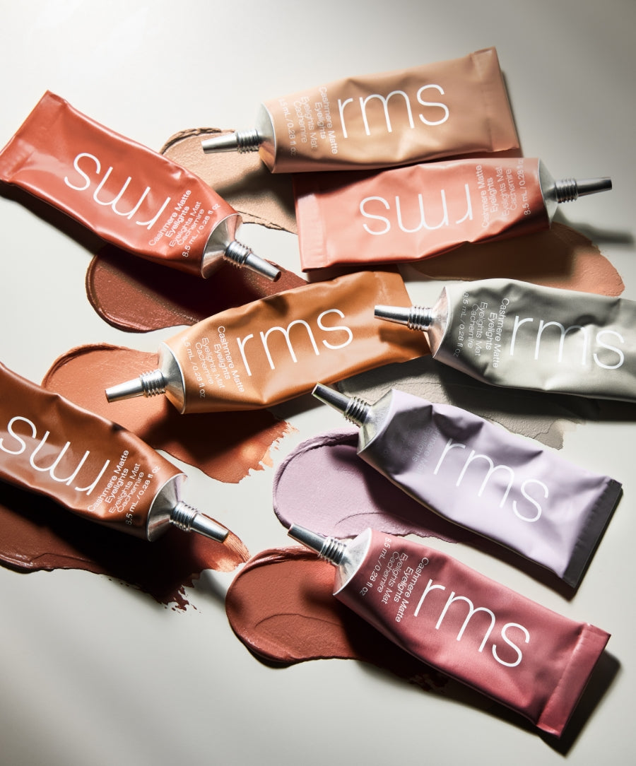 Seven tubes of RMS Beauty Cashmere Matte Eyelights makeup in various soft, natural shades are displayed on a light surface with swatches of each color spread beneath them, highlighting clean beauty, natural ingredients, and sustainable packaging.