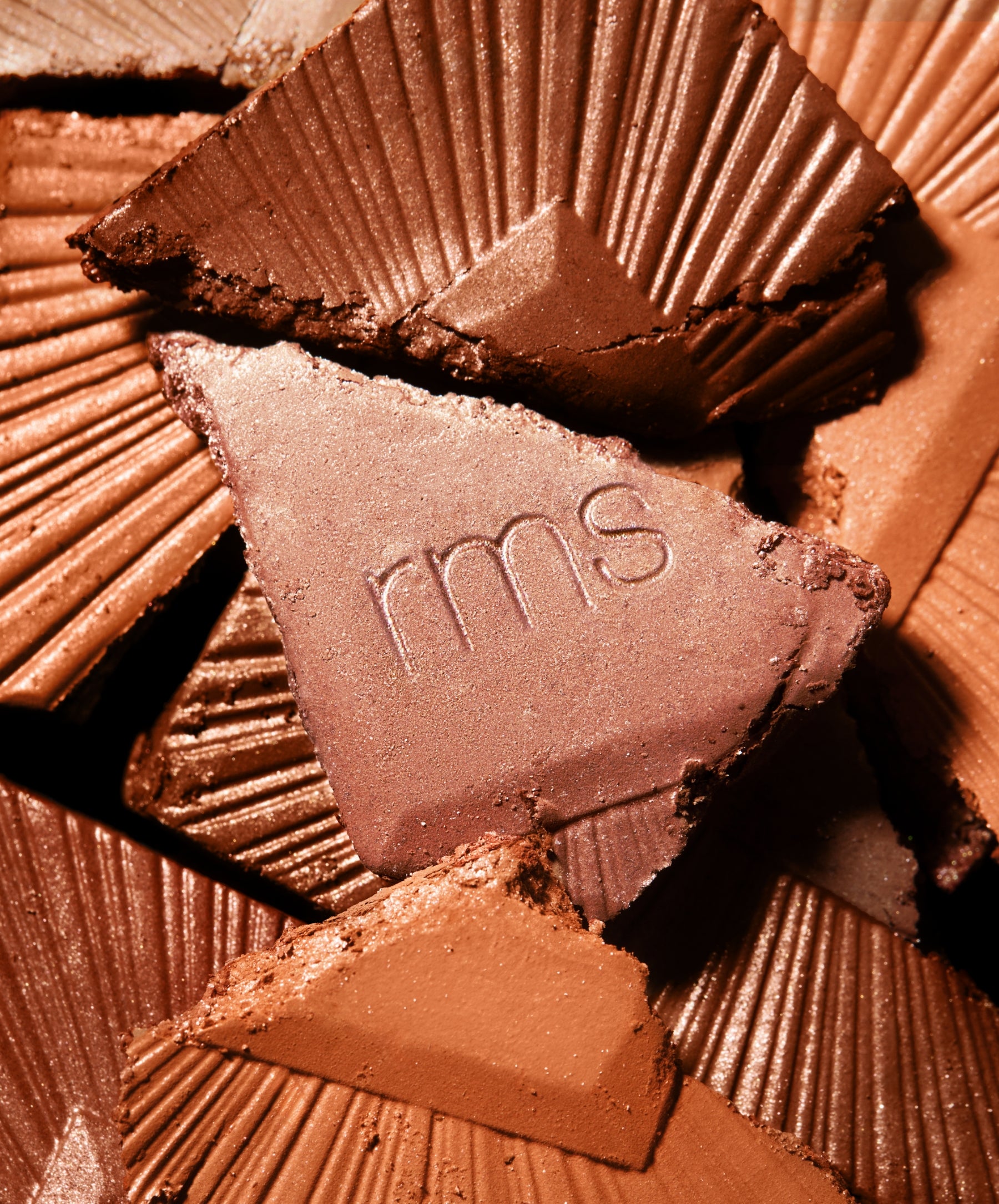 Close-up of broken ReDimension Hydra Bronzer pieces in warm brown shades with embossed "rms" logo on one piece, highlighting smooth, creamy texture and natural ingredients.