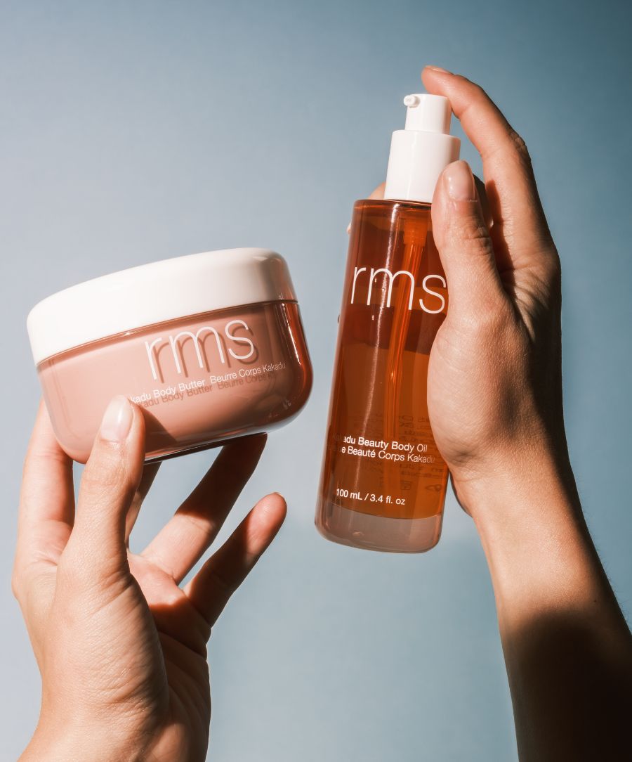 Hands holding a jar and a pump bottle of RMS Beauty products against a light blue background, featuring The Radiant Body Duo for clean beauty and natural body care.