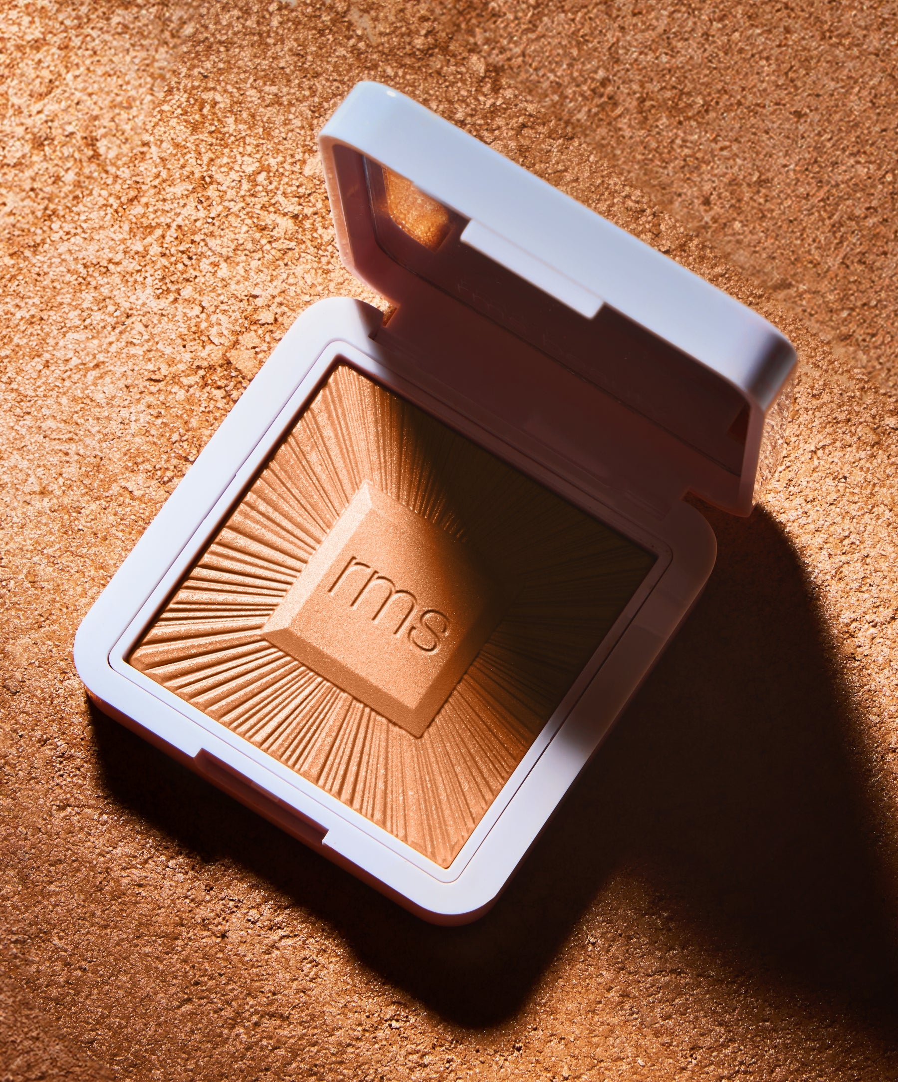 Open compact of ReDimension Hydra Bronzer with a sunburst embossed design, placed on a textured bronze surface, highlighting its warm, natural bronze shade and clean beauty appeal.