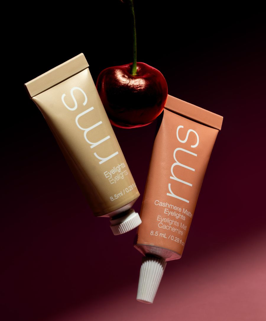 Two tubes of RMS Beauty Cashmere Matte Eyelights in neutral tones placed near a shiny cherry against a dark background, highlighting clean beauty and natural ingredients.