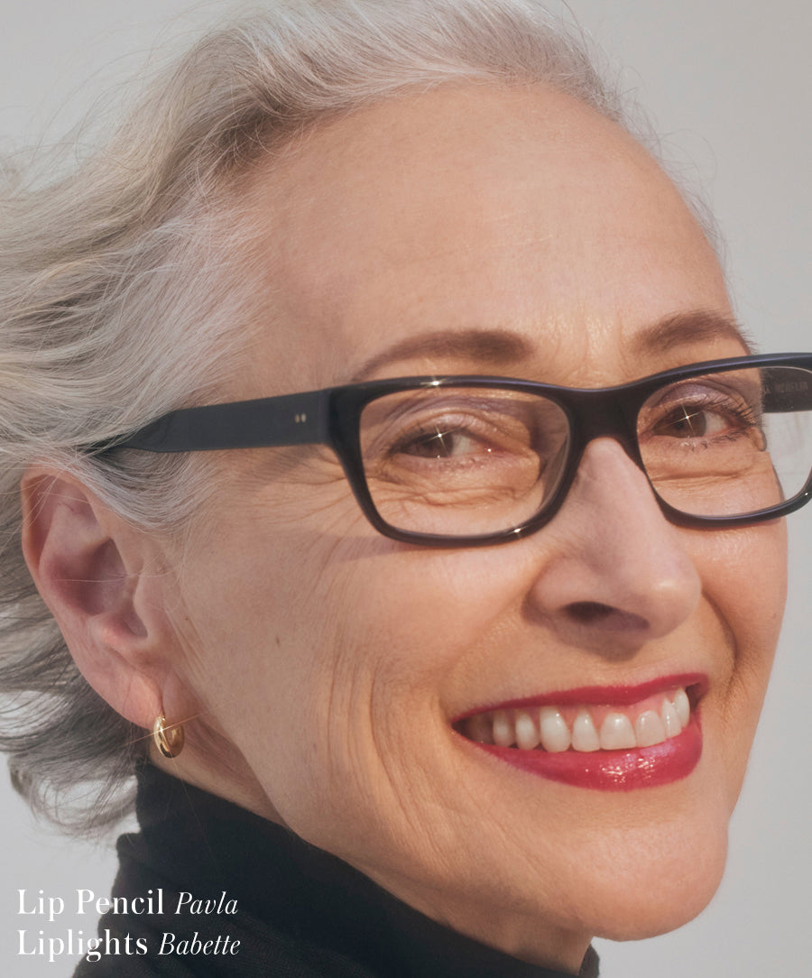 Close-up of an older woman wearing black glasses and gold hoop earrings, smiling with glossy, vibrant lips enhanced by Liplights Cream Lip Gloss, showcasing clean, natural beauty.
