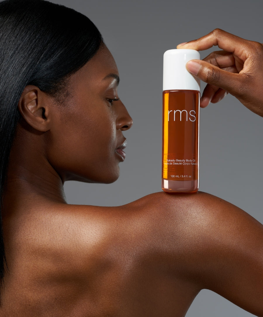 A close-up of a woman with smooth skin holding a bottle of Kakadu Body Oil by RMS Beauty against her shoulder, highlighting natural body care and clean beauty.