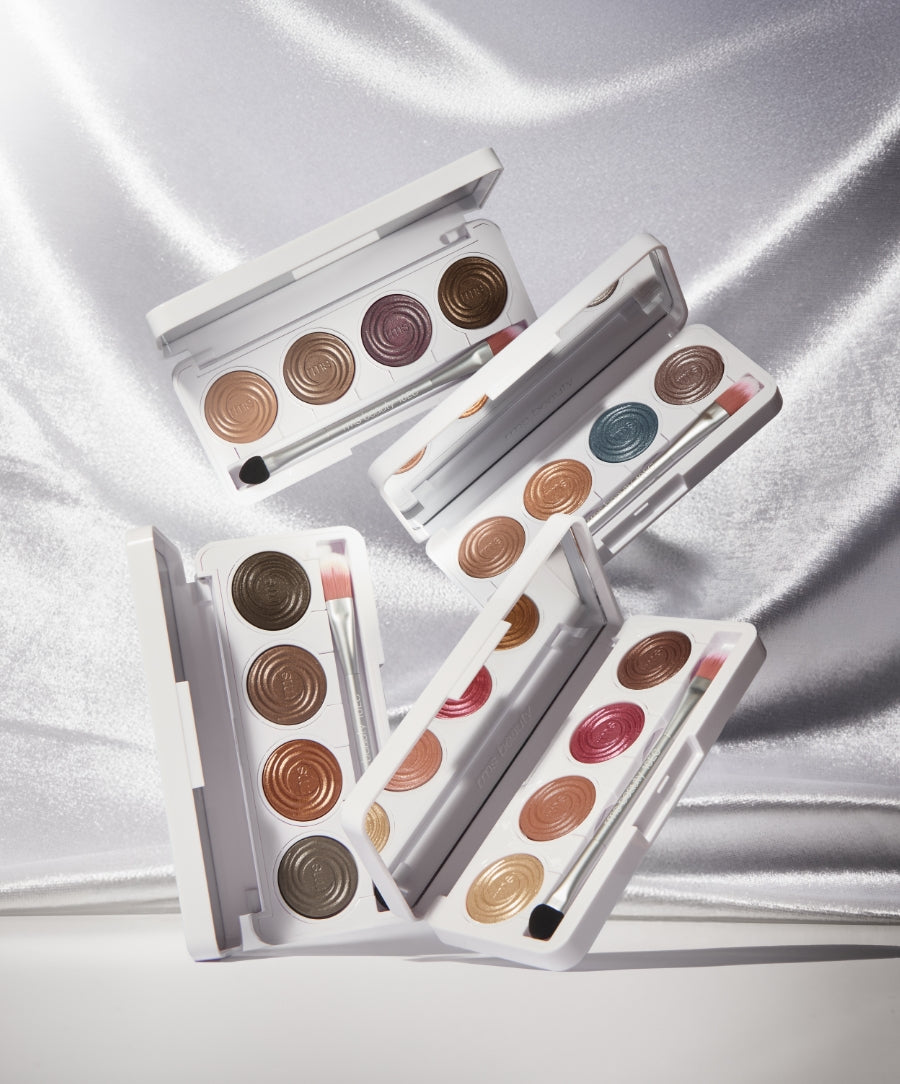 Four open ReDimension Hydra Eyes Quartet palettes showcasing a variety of creamy neutral and jewel-toned eyeshadows with applicator brushes on a shimmering silver fabric background.