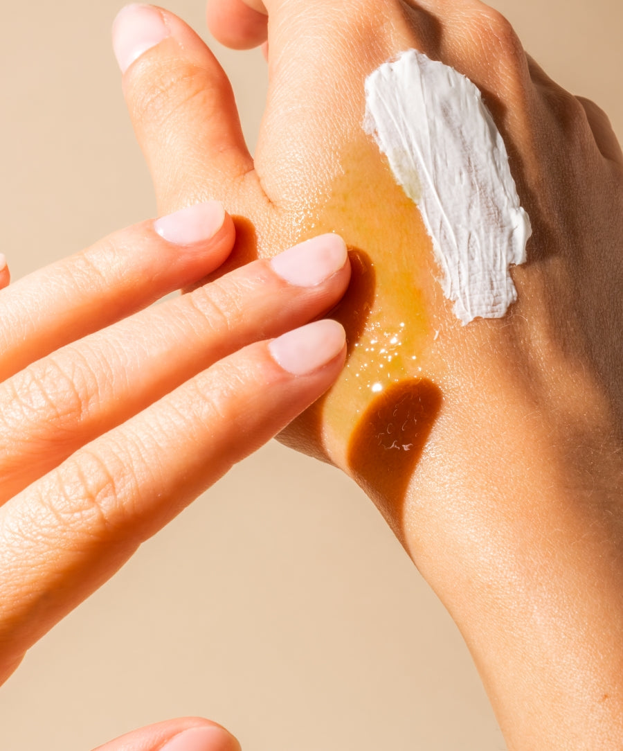 Close-up of a hand with smooth skin applying two textures of skincare products, a creamy white balm and a golden oil, demonstrating moisturizing benefits of The Radiant Body Duo.