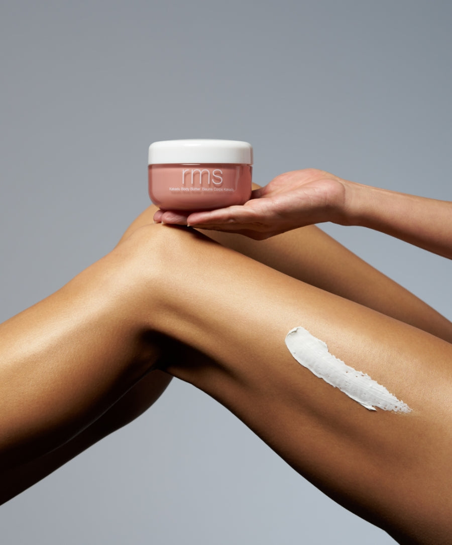 Hand holding a jar of Kakadu Body Butter above smooth, tan legs with a dollop of white cream applied on one leg, set against a neutral gray background.