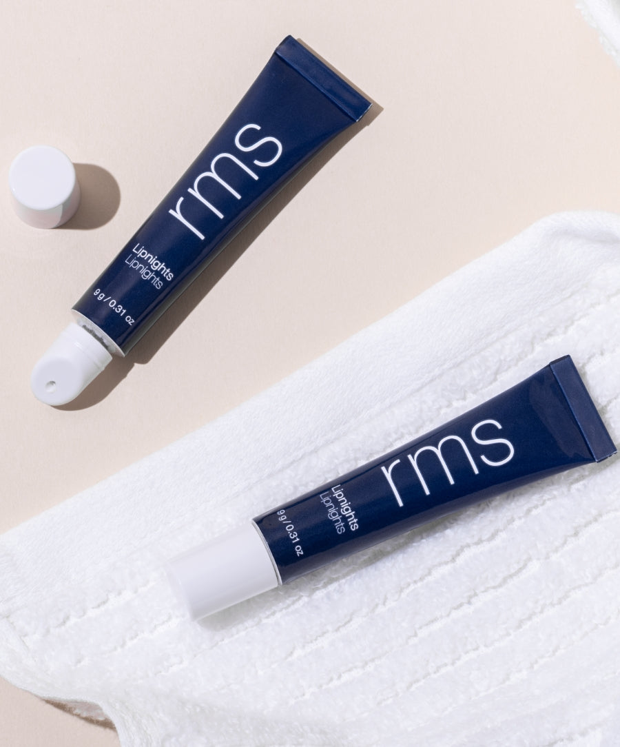 Two navy blue tubes of Lipnights Overnight Lip Mask with white caps, one open and one closed, displayed on a soft white textured cloth against a light background.