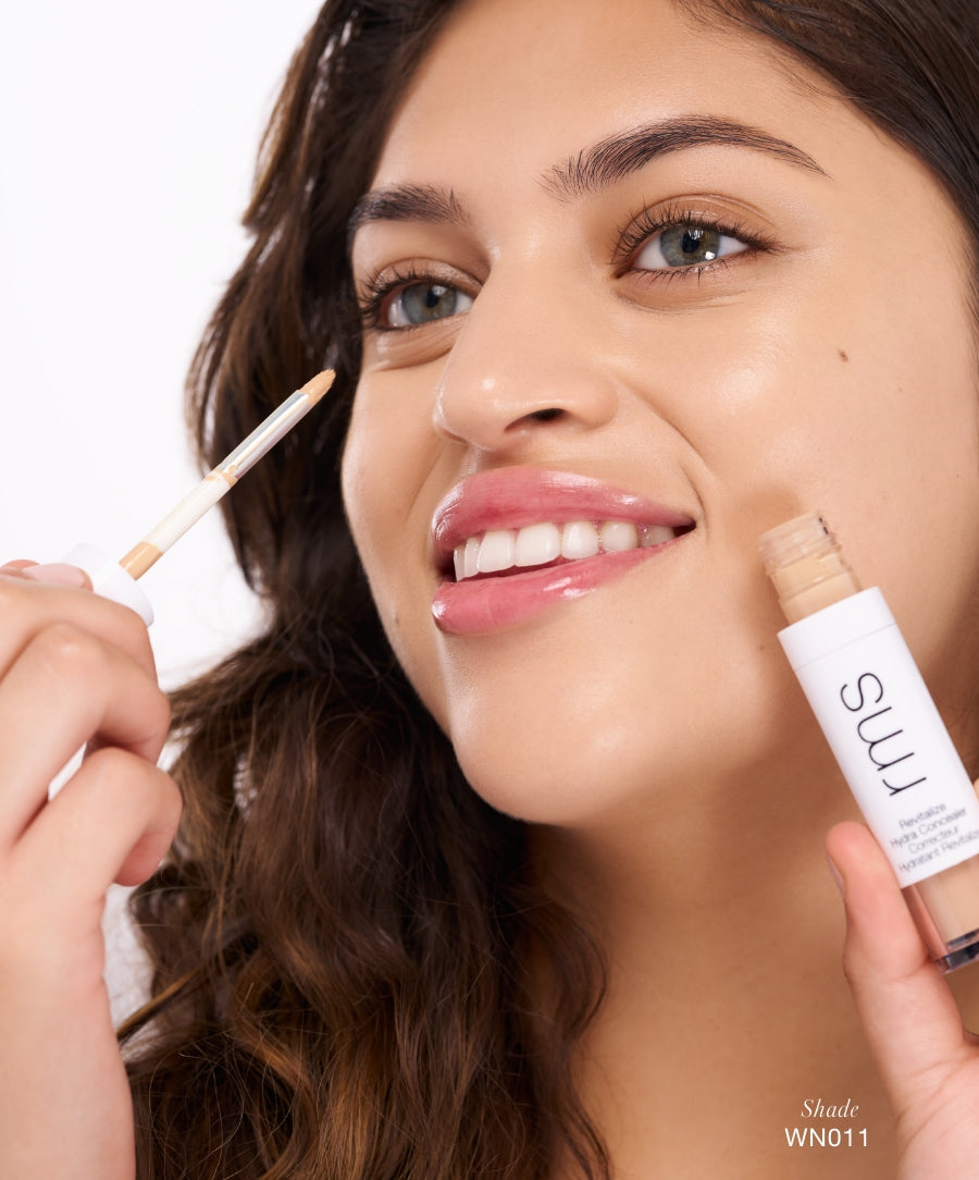 A smiling woman with clear skin holds and applies Revitalize Hydra Concealer with a brush applicator near her eye, showcasing natural beauty and skincare-infused makeup.