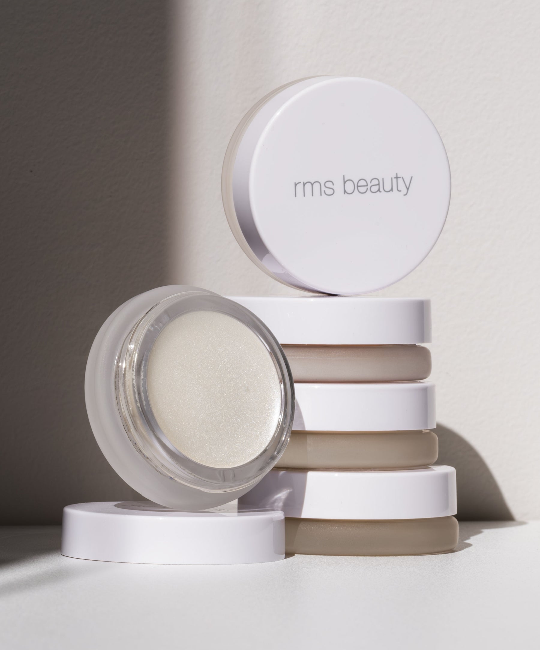 Stack of white RMS Beauty containers with one open jar showing a shimmering cream product, highlighting the Luminizer with clean beauty and natural ingredients.