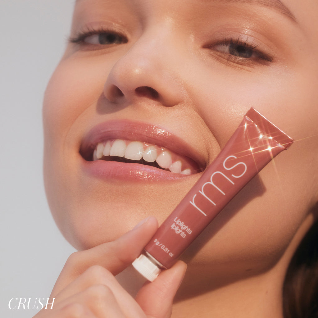 Close-up of a smiling woman holding a tube of Liplights Cream Lip Gloss in a natural, warm shade, showcasing clean beauty and radiant, glossy lips.
