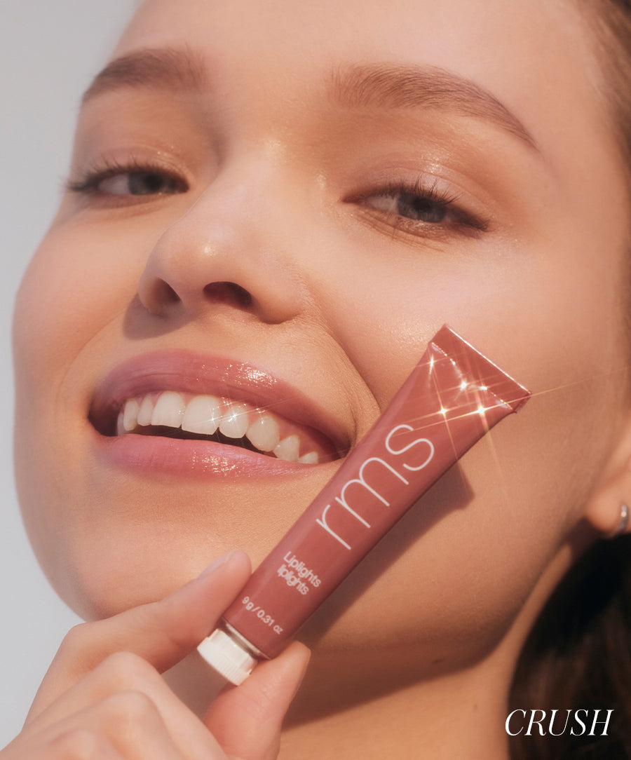 Close-up of a smiling woman holding a tube of Liplights Cream Lip Gloss with natural makeup, showcasing glossy, radiant lips and clean beauty appeal.