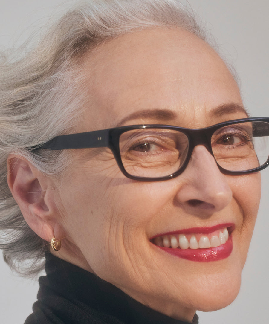 Close-up of a mature woman smiling wearing black glasses and glossy red lips featuring Liplights Cream Lip Gloss, showcasing a radiant and natural makeup look.